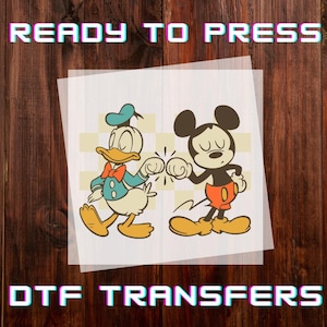 May include: DTF transfer featuring Donald Duck and Mickey Mouse bumping fists. Donald Duck is wearing a blue sailor suit and Mickey Mouse is wearing red shorts. The words "READY TO PRESS" and "DTF TRANSFERS" are displayed in the image.