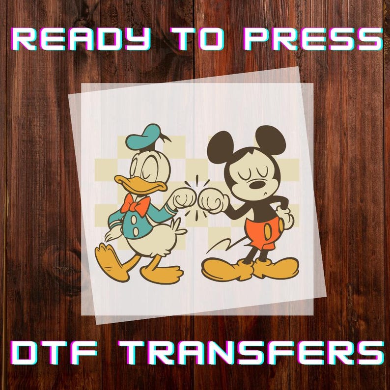 May include: DTF transfer featuring Donald Duck and Mickey Mouse bumping fists. Donald Duck is wearing a blue sailor suit and Mickey Mouse is wearing red shorts. The words "READY TO PRESS" and "DTF TRANSFERS" are displayed in the image.