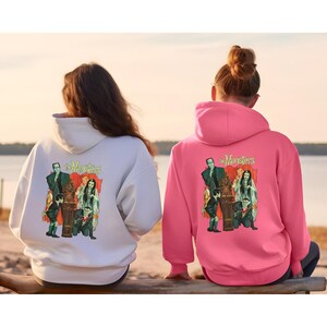 Peut inclure: Deux sweats &agrave; capuche, un blanc et un rose, avec un graphisme vintage de la famille Munsters. Le graphisme comprend le texte "The Munsters" et des images des personnages. Les sweats &agrave; capuche ont un design classique.