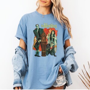 Peut inclure: T-shirt bleu clair avec un motif vintage de la famille Munster. Le design comprend des personnages comme Herman Munster, Lily Munster et Eddie Munster, avec le logo de l'&eacute;mission en haut. Le t-shirt a des manches courtes et une coupe d&eacute;contract&eacute;e.