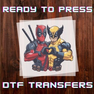May include: DTF transfer featuring Deadpool and Wolverine making a heart shape with their hands. Deadpool is in a red and black suit with swords on his back. Wolverine is in a yellow and blue suit. The words "READY TO PRESS" and "DTF TRANSFERS" are at the top and bottom.