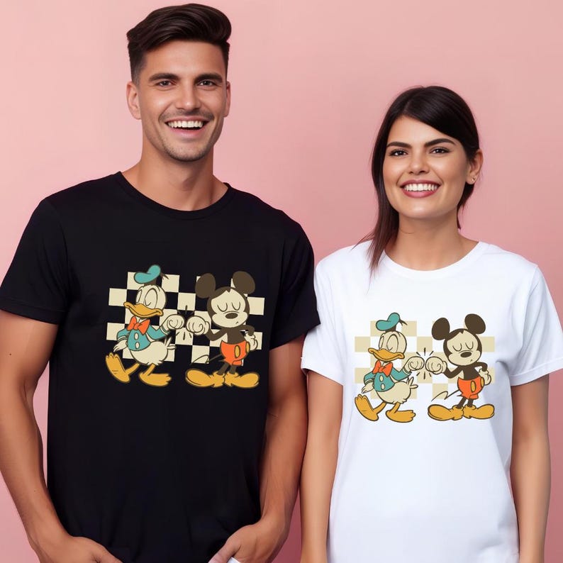 May include: Black and white t-shirts featuring a cartoon graphic of Donald Duck and Mickey Mouse. The characters are set against a checkered background. The t-shirts are worn by two people, one wearing a black shirt and the other a white shirt.