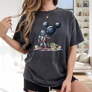 May include: Dark gray t-shirt featuring a graphic of The Mandalorian holding a black balloon shaped like Mickey Mouse ears, with Grogu standing nearby. The design includes colorful orbs. The shirt is a casual, unisex style.