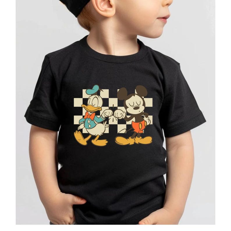 May include: Black t-shirt featuring Donald Duck and Mickey Mouse. Donald Duck is wearing a blue bow tie and hat, and Mickey Mouse is wearing red shorts. The characters are set against a black and white checkered background.