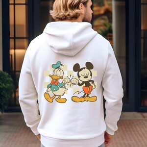 May include: White hoodie featuring a graphic of Donald Duck and Mickey Mouse bumping fists. The cartoon characters are set against a yellow and white checkered background. The hoodie is a casual, comfortable garment.