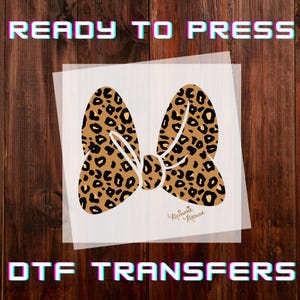 May include: A DTF transfer featuring a leopard print bow design with the text "READY TO PRESS" and "DTF TRANSFERS". The bow has a tan background with black spots and a white outline. The words "Minnie Mouse" are written in cursive.