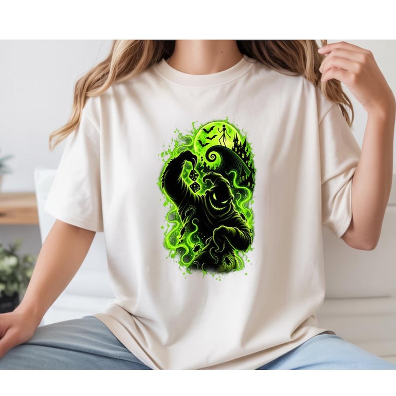 May include: A cream-colored t-shirt featuring a graphic print of a black, sack-like character holding dice. The design includes a bright green glowing background with silhouettes of a crooked house, a full moon, bats, and small spiders.