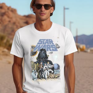 May include: White t-shirt with a Star Wars graphic. The design features Darth Vader, other characters, and the Star Wars logo in blue and yellow. The shirt has a classic crew neck.