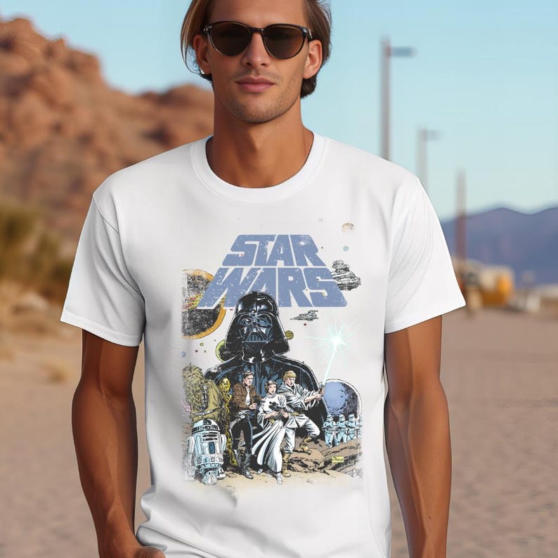May include: White t-shirt with a Star Wars graphic. The design features Darth Vader, other characters, and the Star Wars logo in blue and yellow. The shirt has a classic crew neck.