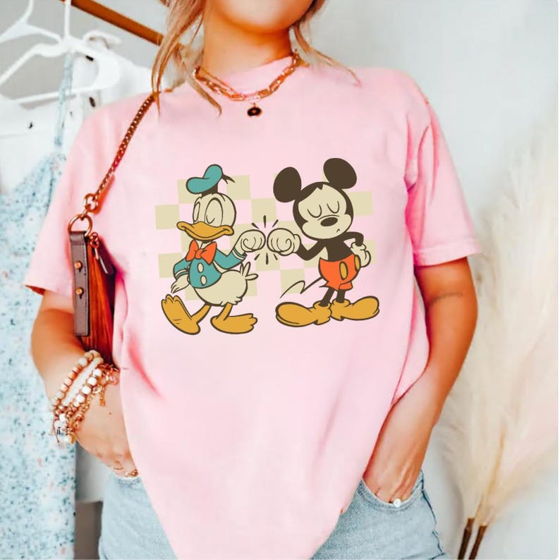 May include: Pink t-shirt featuring a graphic of Donald Duck and Mickey Mouse bumping fists. Donald Duck is wearing a blue jacket and hat, while Mickey Mouse is in red shorts. The background has a checkered pattern.