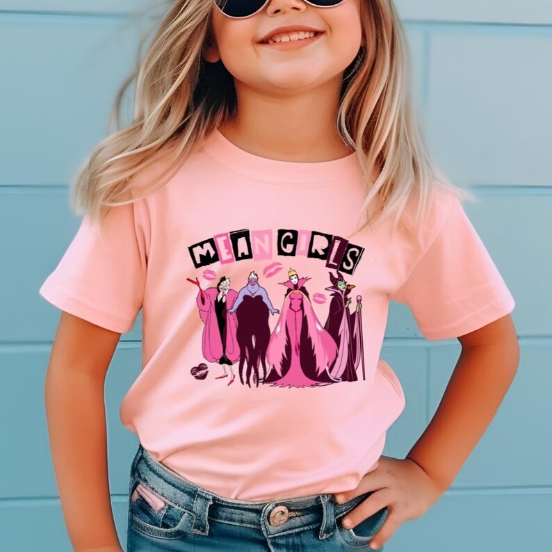 May include: A pale pink t-shirt with the words "MEAN GIRLS" and a graphic of cartoon villains in pink and purple. The t-shirt is worn by a person with blonde hair and sunglasses.