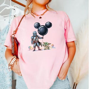 May include: Pink t-shirt featuring a graphic of The Mandalorian and Grogu, with a black Mickey Mouse balloon. The Mandalorian is in full armor, holding the balloon, and Grogu stands beside him. Colorful orbs are in the background.