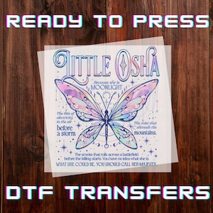 May include: A DTF transfer sheet with the text "READY TO PRESS" and "DTF TRANSFERS". The design features a butterfly with iridescent wings and the words "LITTLE OSHA" and other text. The background is a wooden surface.