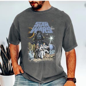 May include: Grey t-shirt with a Star Wars graphic. The design features the Star Wars logo, Darth Vader, and other characters from the film. The shirt is a short-sleeved crew neck.