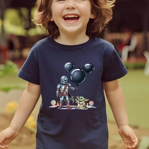May include: Navy blue t-shirt featuring a cartoon illustration of The Mandalorian holding a black balloon with Mickey Mouse ears, and Grogu. The design includes colorful accents and is suitable for children.