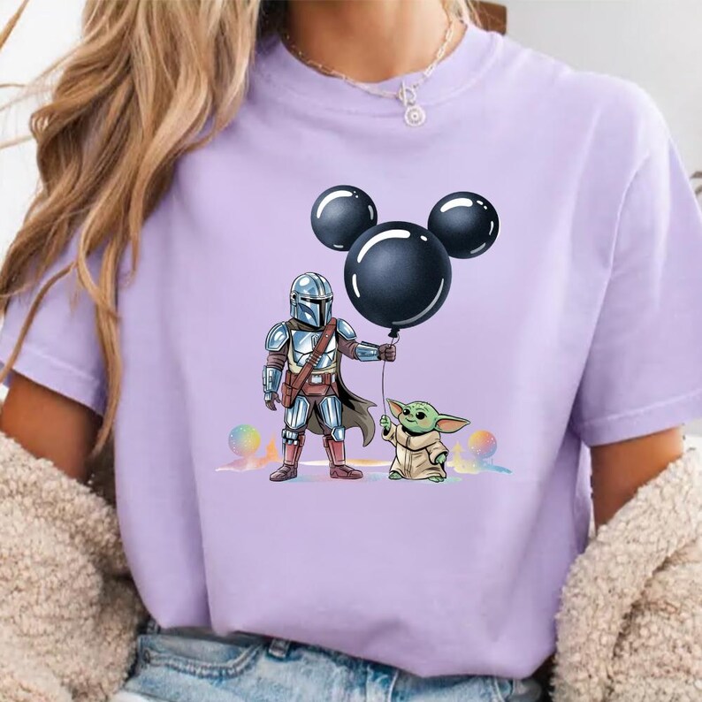 May include: A light purple t-shirt featuring a graphic of The Mandalorian and Grogu holding a black balloon. The Mandalorian is in silver armor, and Grogu is in a tan cloak. The balloon has a Mickey Mouse head shape.