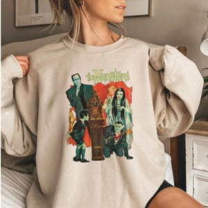Peut inclure: Sweat-shirt beige avec un graphisme color&eacute; de la famille Munster. Le graphisme comprend les mots "The Munsters" en vert, avec des images des personnages dans leurs tenues classiques. Le sweat-shirt est de couleur neutre, adapt&eacute; aux tenues d&eacute;contract&eacute;es.