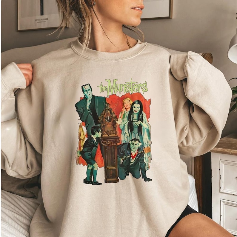 Peut inclure: Sweat-shirt beige avec un graphisme color&eacute; de la famille Munster. Le graphisme comprend les mots "The Munsters" en vert, avec des images des personnages dans leurs tenues classiques. Le sweat-shirt est de couleur neutre, adapt&eacute; aux tenues d&eacute;contract&eacute;es.