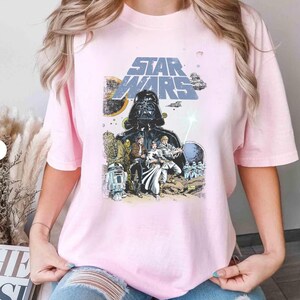 May include: A pale pink t-shirt with a Star Wars graphic. The design features Darth Vader, various characters, and the Star Wars logo in blue and white. The shirt has short sleeves and a crew neck.