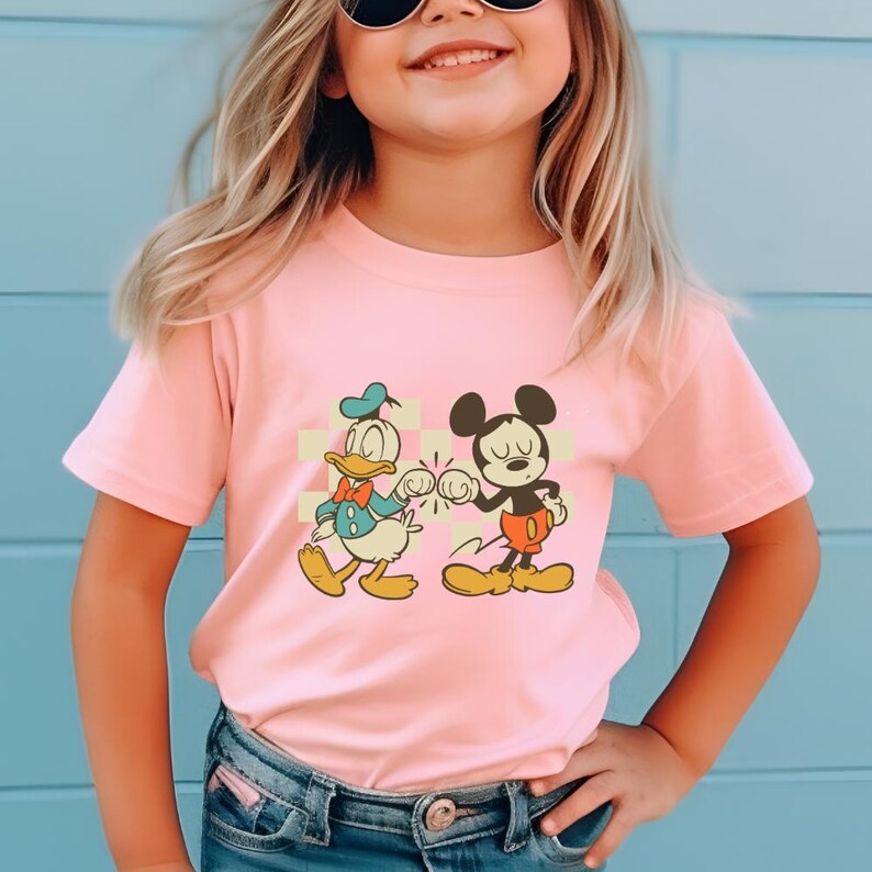 May include: A light pink t-shirt featuring a graphic of Donald Duck and Mickey Mouse bumping fists. The cartoon characters are in a vintage style, with Donald Duck wearing a blue sailor suit and Mickey Mouse in red shorts. The shirt is worn by a child.