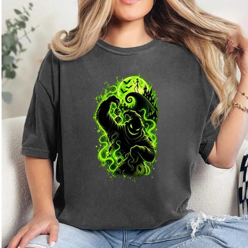 May include: A charcoal gray short-sleeve t-shirt featuring a vibrant neon green graphic print of a sack-like character holding dice, surrounded by swirling smoke, bats, a crescent moon, and a silhouette of a crooked house on a hill.