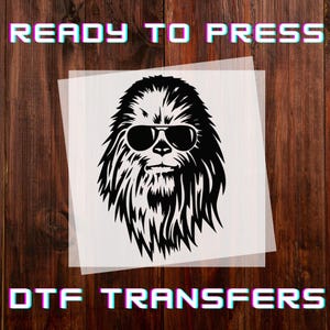 May include: Black and white DTF transfer featuring a stylized image of a Wookiee wearing sunglasses. The words "READY TO PRESS" and "DTF TRANSFERS" are displayed in a retro font. The transfer is on a transparent sheet, set against a wooden background.