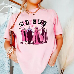 May include: A pale pink t-shirt with the words "MEAN GIRLS" above a cartoon graphic of four villains in pink and purple. The t-shirt has a crew neck and short sleeves.