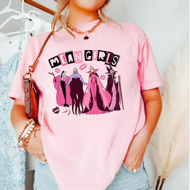May include: A pale pink t-shirt with the words "MEAN GIRLS" above a cartoon graphic of four villains in pink and purple. The t-shirt has a crew neck and short sleeves.