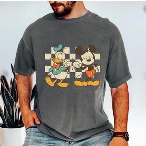 May include: Dark gray t-shirt featuring a graphic of Donald Duck and Mickey Mouse. The cartoon characters are standing in front of a checkered background. Donald Duck is wearing a blue jacket and Mickey Mouse is wearing red shorts.