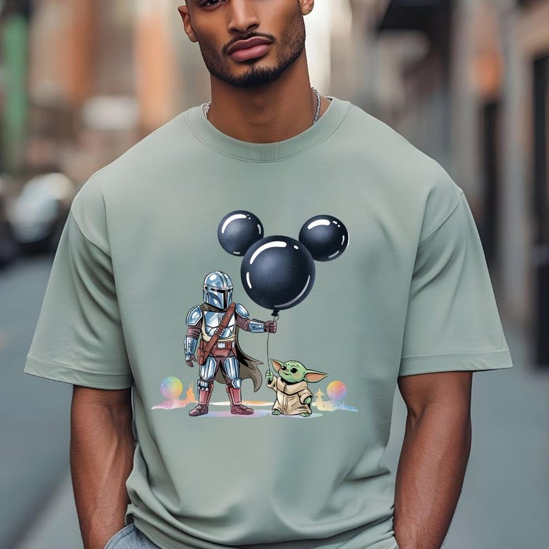 May include: Sage green t-shirt featuring a graphic of The Mandalorian holding a black balloon with Mickey Mouse ears, and Grogu. The design includes colorful accents. The shirt is a casual wear item.