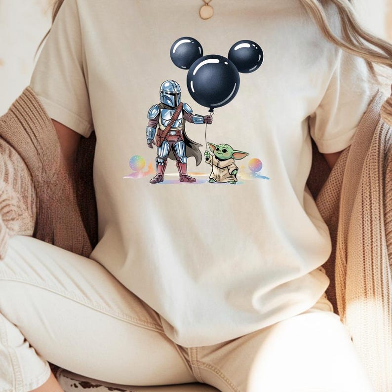 May include: Beige t-shirt featuring a graphic of The Mandalorian and Grogu holding a black Mickey Mouse balloon. The Mandalorian is in full armor, and Grogu is in a tan robe. Colorful orbs are in the background.