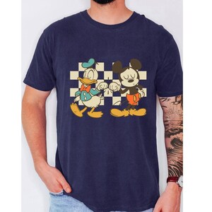 May include: Navy blue t-shirt featuring a retro graphic of Donald Duck and Mickey Mouse fist-bumping in front of a checkered background. Donald Duck is wearing a blue outfit and Mickey Mouse is wearing red shorts. The shirt is a classic crew neck.