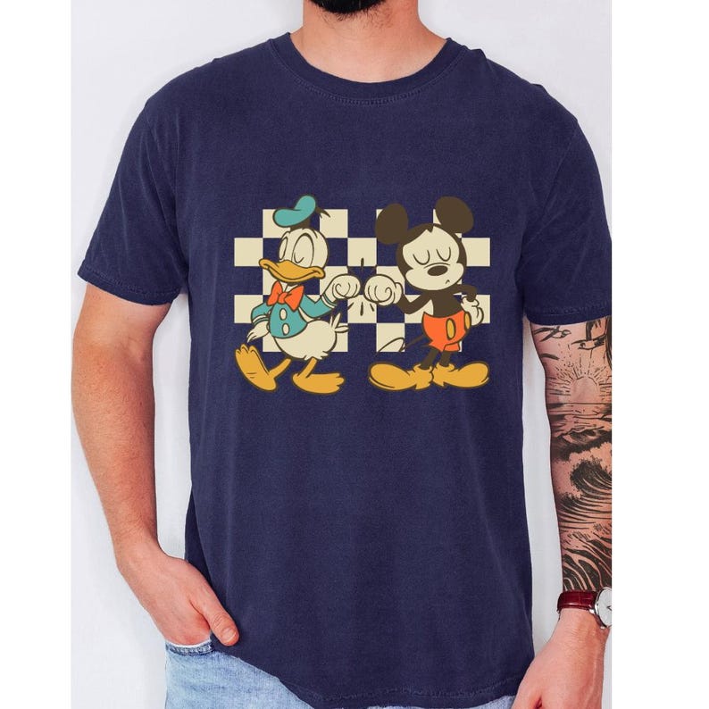 May include: Navy blue t-shirt featuring a retro graphic of Donald Duck and Mickey Mouse fist-bumping in front of a checkered background. Donald Duck is wearing a blue outfit and Mickey Mouse is wearing red shorts. The shirt is a classic crew neck.