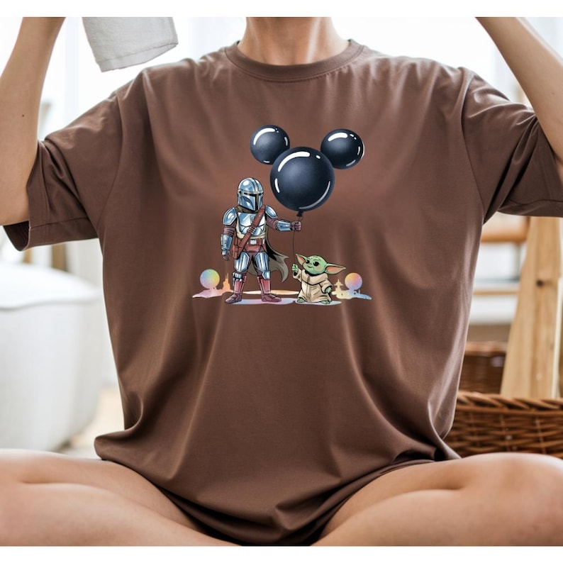 May include: Brown t-shirt featuring a graphic of The Mandalorian and Grogu, with a black Mickey Mouse balloon. The illustration includes colorful accents and details. The shirt is a casual wear item.