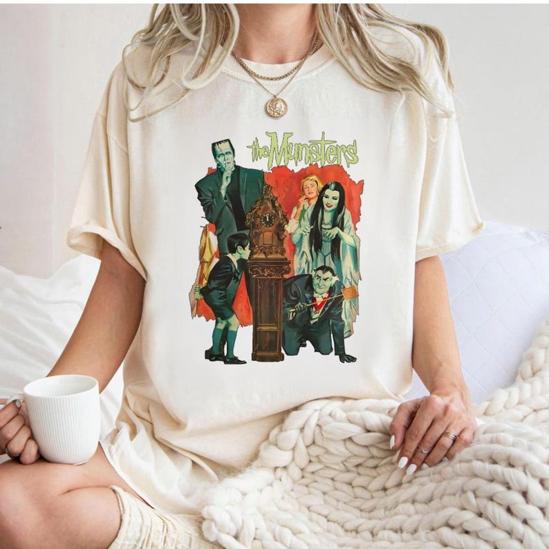 Peut inclure: T-shirt cr&egrave;me avec un graphisme vintage de la famille The Munsters. Le design comprend les personnages dans leurs tenues classiques, avec le logo de l'&eacute;mission en vert. L'&oelig;uvre est plac&eacute;e sur un fond rouge.