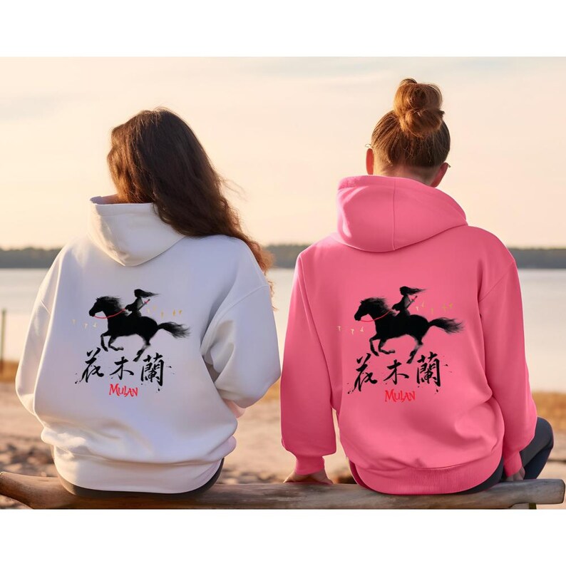 Pode incluir: Duas camisolas com capuz, uma branca e outra rosa, apresentam um gr&aacute;fico preto de uma pessoa montada a cavalo, com caracteres chineses e a palavra "Mulan" em vermelho. As camisolas t&ecirc;m capuz com cord&atilde;o e um bolso canguru.
