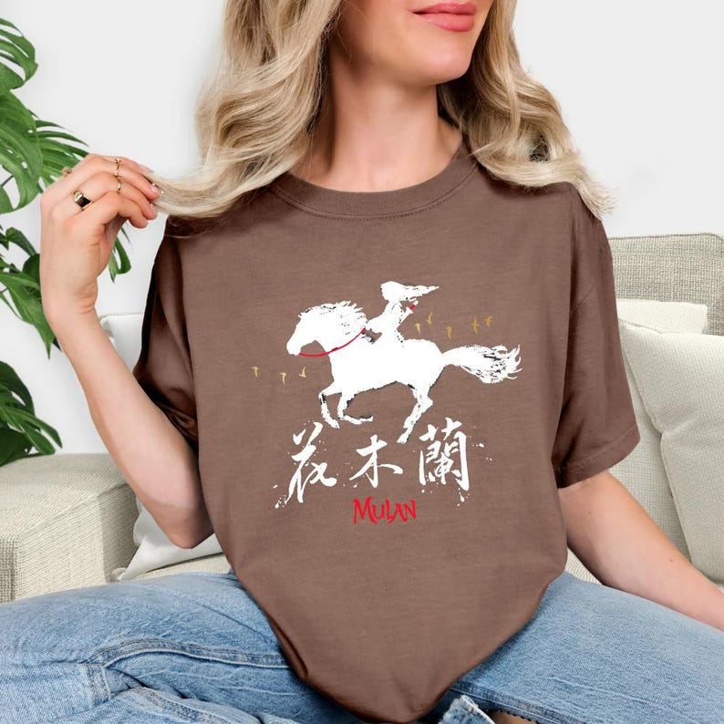 Pode incluir: Camiseta marrom com um gr&aacute;fico branco de uma pessoa montando um cavalo, caracteres chineses e a palavra "MULAN" em vermelho. O design inclui pequenos p&aacute;ssaros. A camiseta &eacute; feita de material macio.