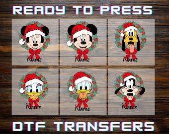 May include: Six DTF transfers featuring cartoon characters wearing Santa hats, each framed by a green Christmas wreath with a red bow and the word Name below. The text READY TO PRESS DTF TRANSFERS appears at the top and bottom of the image.