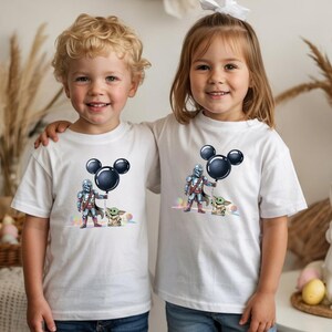 May include: Two children wearing white t-shirts with a graphic featuring a character in silver armor holding a black Mickey Mouse balloon, with a small green creature. The shirts are a light color, and the graphic is colorful.