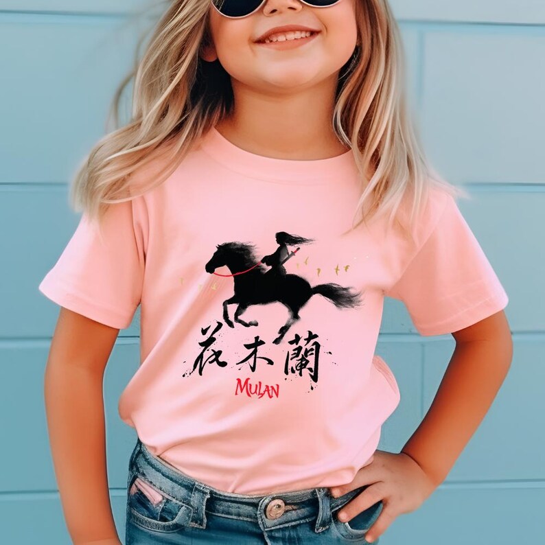 Pode incluir: T-shirt rosa claro com uma silhueta preta de uma pessoa montando um cavalo. A camisa tem caracteres chineses e a palavra "Mulan" em vermelho. A camisa &eacute; usada por uma crian&ccedil;a.
