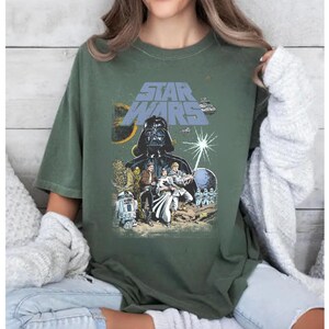 May include: Olive green Star Wars graphic t-shirt featuring Darth Vader, Luke Skywalker, Princess Leia, and other characters. The Star Wars logo is in blue. The shirt has a vintage, distressed look.