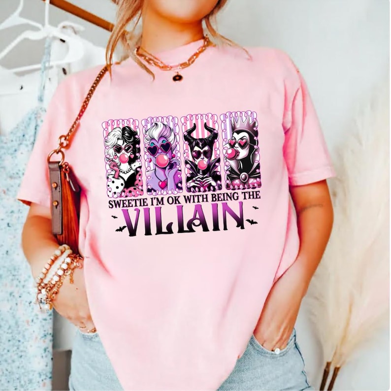 May include: Pink t-shirt featuring a graphic of four cartoon villains blowing bubblegum bubbles. The text "Sweetie I'm OK with being the Villain" is printed below the characters in black. The shirt is a light pink color.