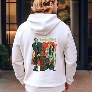 Peut inclure: Sweat &agrave; capuche blanc avec un graphisme color&eacute; de la famille Munster et le texte "The Munsters" au dos. Le graphisme comprend Frankenstein, un vampire et d'autres personnages de la s&eacute;rie t&eacute;l&eacute;vis&eacute;e. Le sweat &agrave; capuche a une capuche &agrave; cordon et une poche kangourou.