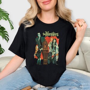 Peut inclure: T-shirt noir avec un graphisme color&eacute; de la famille Munster. Le graphisme comprend des images d'Herman, Lily, Grandpa, Eddie et Marilyn Munster, avec le texte "The Munsters" en vert et jaune.