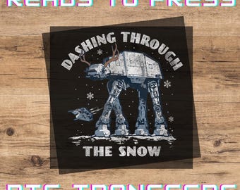 May include: A square DTF transfer featuring a grey AT-AT walker with reindeer antlers, surrounded by white snowflakes and a small flying ship. The text reads DASHING THROUGH THE SNOW in a distressed white font. The words READY TO PRESS and DTF TRANSFERS appear in large, bold, cyan-outlined letters at the top and bottom.