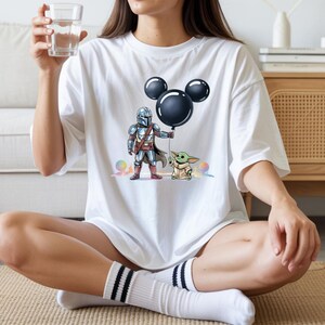May include: White t-shirt featuring a cartoon illustration of a Mandalorian character and Grogu holding a black Mickey Mouse balloon. The design includes colorful accents and is printed on a short-sleeved shirt.