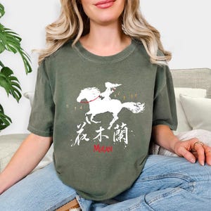 Pode incluir: Camiseta verde-oliva com um gr&aacute;fico branco de uma pessoa montando um cavalo, com o texto "花木蘭" e "Mulan" em vermelho. O design &eacute; uma ilustra&ccedil;&atilde;o estilizada. A camiseta tem mangas curtas e gola redonda.