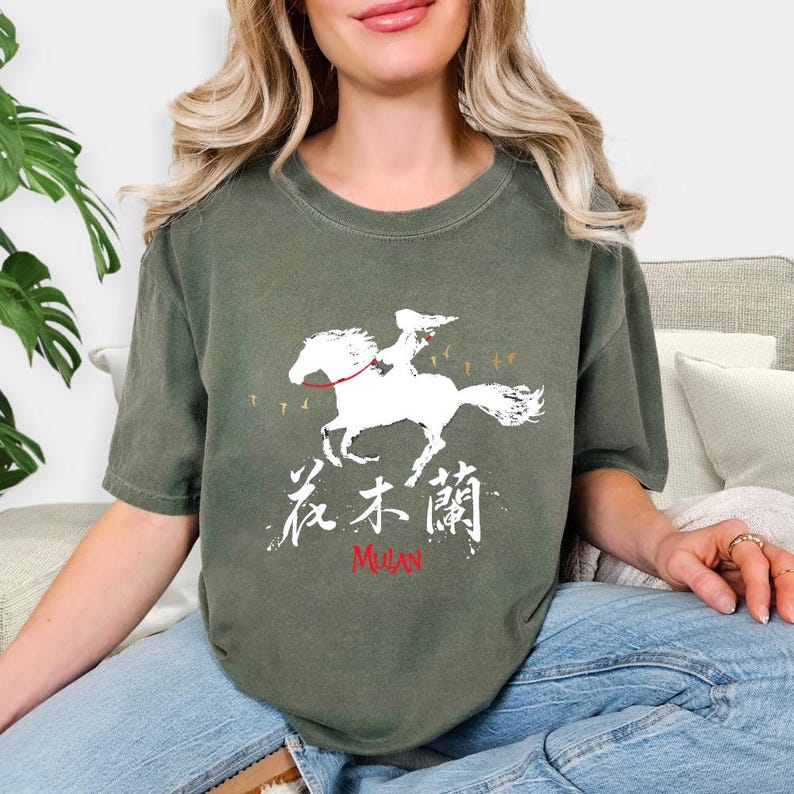 Pode incluir: Camiseta verde-oliva com um gr&aacute;fico branco de uma pessoa montando um cavalo, com o texto "花木蘭" e "Mulan" em vermelho. O design &eacute; uma ilustra&ccedil;&atilde;o estilizada. A camiseta tem mangas curtas e gola redonda.