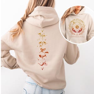 Op de afbeelding: Beige hoodie met een drakenontwerp op de rug in tinten oranje en rood. Het ontwerp toont een reeks gestileerde draken. Een kleinere afbeelding op de voorkant toont een cirkelvormig ontwerp met de woorden "Iron Flame" en een decoratieve rand.