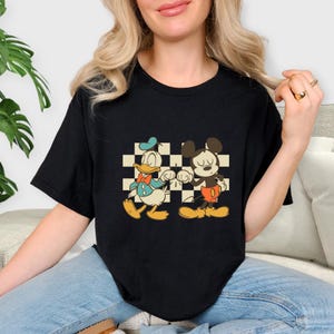 May include: Black t-shirt featuring a cartoon graphic of Donald Duck and Mickey Mouse holding hands in front of a checkered background. Donald Duck is wearing a blue hat and Mickey Mouse is wearing red shorts.